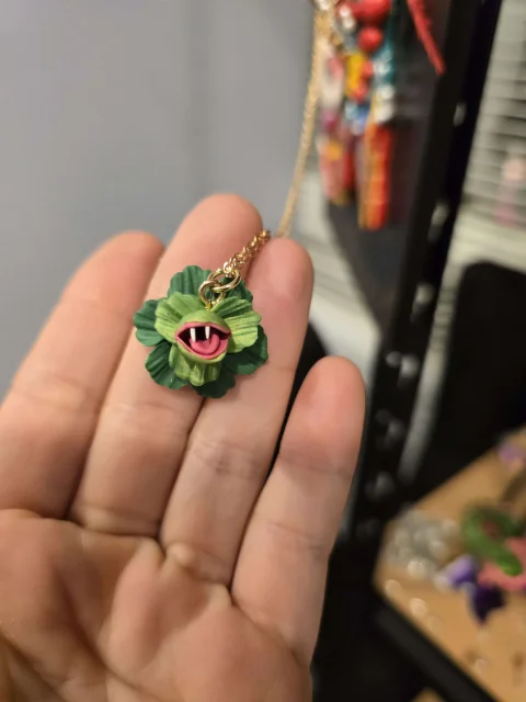 A tiny green, one-eyed sculpted creature with an open red mouth, resting on the maker’s hand.