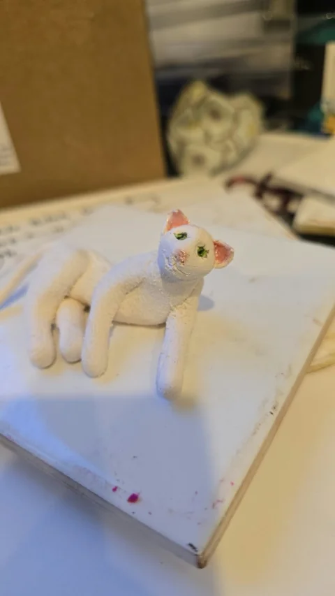 A small pale clay cat with colorful ears and a smiling face, standing on a worktable.